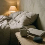 A peaceful bedside still life with a ceramic tea mug, closed linen book, and river stone beside linen bedding in soft evening light